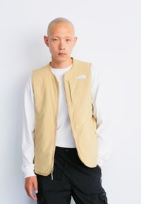 Lightweight, sleeveless beige vest with a smooth texture and zip closure. Features a small white logo on the upper left side.