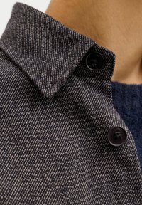 Tumehall tekstuuriga kangas diagonaalse koega, terava krae ja mustade nööpidega ees. Lähedalt võetud detail rõhutab materjali.