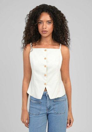 Femme aux cheveux bouclés portant un haut blanc sans manches boutonné et un jean bleu clair avec des poches avant tressées, debout devant un fond gris.