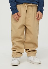 Beżowe bawełniane spodnie z elastycznym pasem i sznurkiem do regulacji. Luźny krój i podwinięte mankiety, w połączeniu z białymi butami sportowymi.