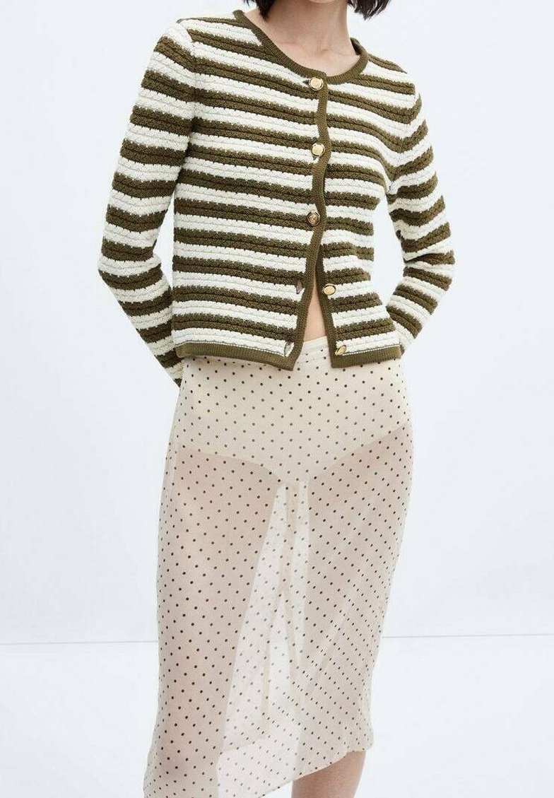 Femme portant un cardigan rayé vert et blanc boutonné avec une jupe beige transparente à pois noirs.