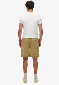 Pantalones cortos de cargo beige con dos bolsillos laterales, cintura elástica y una etiqueta en el bolsillo, combinados con una camiseta blanca lisa y zapatillas beige.