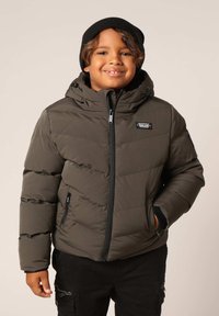 Veste verte matelassée avec fermeture éclair, capuche à cordon de serrage et accents noirs. Portée par-dessus un haut noir et associée à un pantalon sombre.