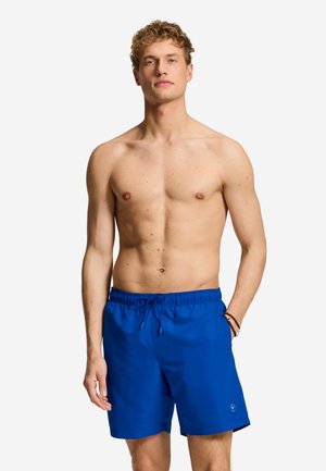 Bărbat fără cămașă cu păr blond ondulat, purtând pantaloni scurți de baie albaștri, stând cu o mână în buzunar pe un fundal simplu.