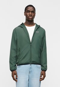 Νέος άνδρας στέκεται φορώντας πράσινο μπουφάν με φερμουάρ και κουκούλα πάνω από λευκό πουκάμισο και ανοιχτόχρωμο τζιν, σε λευκό φόντο.