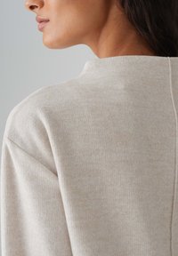 Beige, gerippter Pullover mit hohem Kragen und dezenten Nahtdetails auf der Rückseite, mit weichem Textil und lässiger Silhouette.