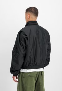 Mees musta bomberjaki ja roheliste sõjapükstega, seistes seljaga kaamera poole tavalise valge tausta ees.