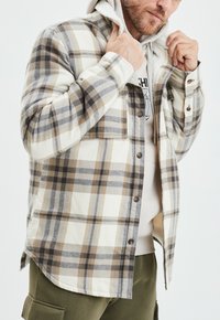 Camicia in flanella a quadri nei colori crema, marrone e grigio. Presenta un morbido rivestimento interno testurizzato, chiusura con bottoni e taschino sul petto.