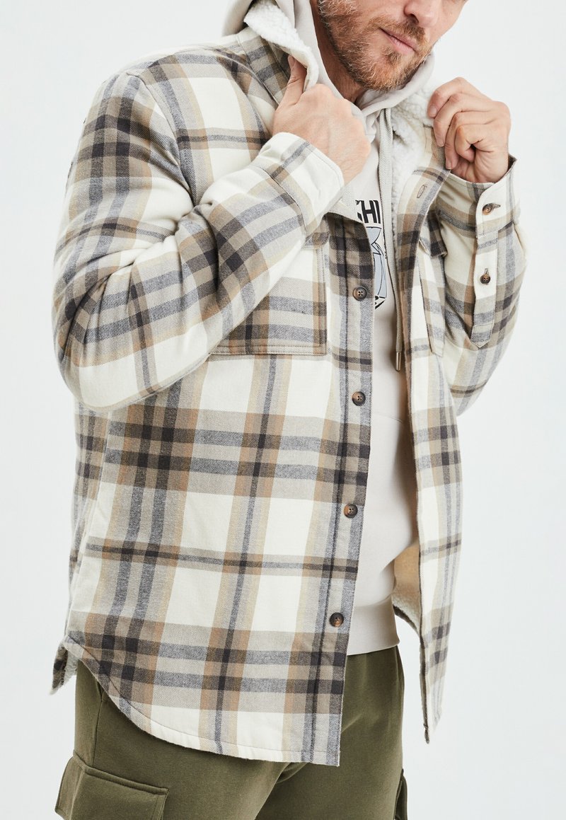 Camicia in flanella a quadri nei colori crema, marrone e grigio. Presenta un morbido rivestimento interno testurizzato, chiusura con bottoni e taschino sul petto.