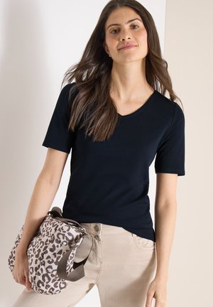 Žena s dlouhými hnědými vlasy v černém tričku s krátkým rukávem a béžových kalhotách, držící po boku tašku s leopardím vzorem.