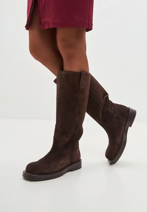 Bottes en suède marron, montant jusqu'au genou, avec une semelle plate et des lignes épurées, présentant des coutures latérales et une construction robuste pour un soutien optimal.