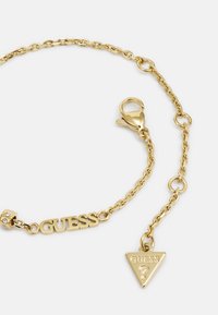 Bracelet en chaîne dorée avec un charm en forme de logo "GUESS" et une étiquette triangulaire avec un symbole de clé; comprend un fermoir en homard et des accents de strass.