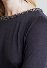 Mörkgrå stickad topp med en strukturerad pärlkrage med metalliska detaljer, rund hals och långa ärmar. Mjuk tyg med subtil lyster.