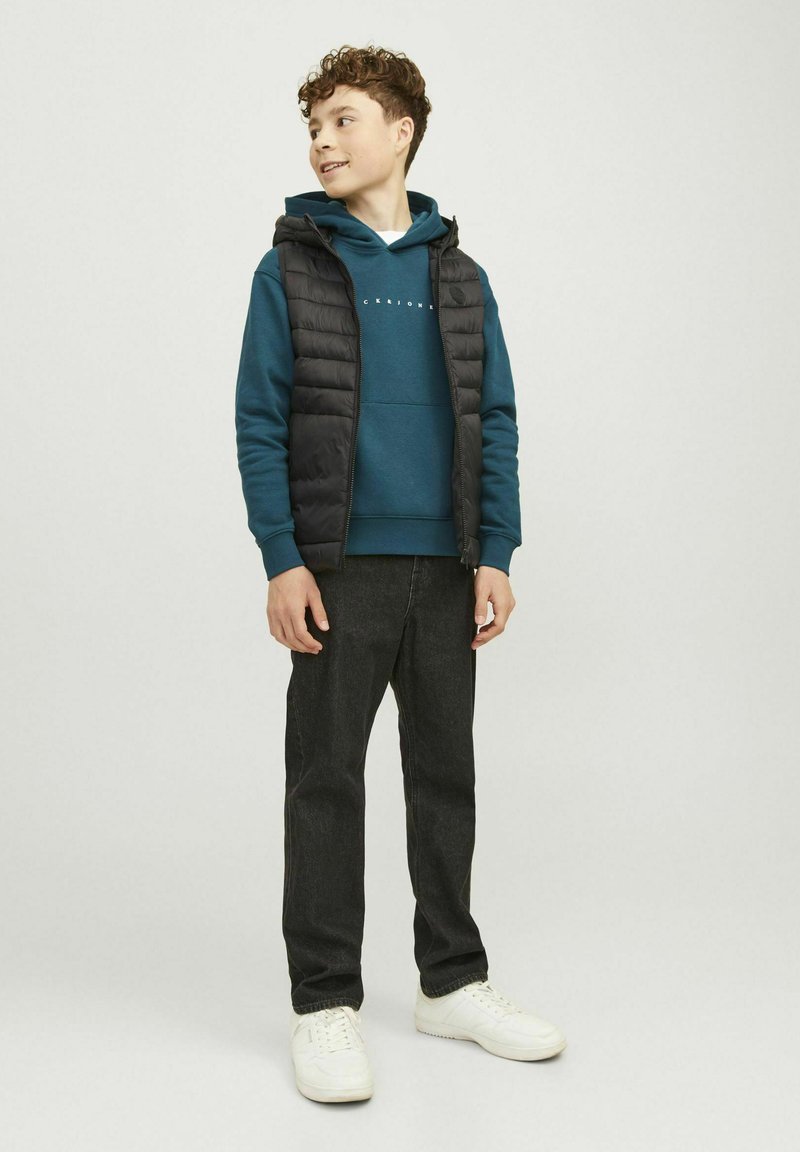 Chaleco acolchado negro sobre sudadera de color teal, combinado con pantalones oscuros y zapatillas blancas. Los detalles incluyen cierre de cremallera y texto impreso en la sudadera.