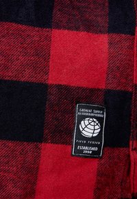 Rood-zwart geruite stof met een gestructureerd patroon. Bevat een zwart label met witte tekst en een wereldbolpictogram. Zacht, warm materiaal.