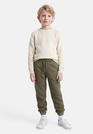 Lyshåret dreng stående, iført en cremefarvet sweater, olivengrønne joggers med snørebånd, hvide sokker og hvide sneakers, foran en ensfarvet baggrund.