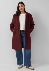 Burgunderfarbener Wollmantel mit Kragen, vorderen Knöpfen und zwei seitlichen Taschen, kombiniert mit blauen Schlagjeans und weißen Sneakers.