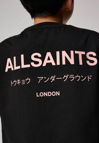 Juoda marškinėliai su dideliu rožiniu tekstu. Tekstas apima "ALLSAINTS," japonų simbolius ir "LONDON." Pagaminti iš medvilnės. Apvalus iškirpimas.
