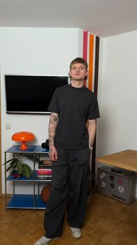 Νεαρός άνδρας με τατουάζ, ντυμένος με μαύρα ρούχα, στέκεται σε ένα δωμάτιο με τηλεόραση επίπεδης οθόνης, πορτοκαλί λάμπα, αθλητική μπάλα και πολύχρωμες ρίγες στον τοίχο.