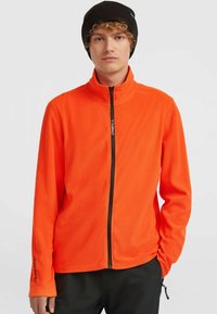 Ung mand med krøllet hår iført en lys orange fleece-jakke med lynlås og en sort beanie, stående med den ene hånd i lommen.