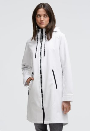 Woman wearing a white hooded coat with black zipper details and drawstrings, standing with one hand in pocket, looking to the side.