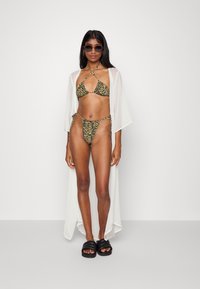 Biquíni com riscas de tigre amarelas e negras, combinado com uma capa branca transparente. Sandálias de slide pretas completam o look.