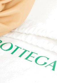 Article en cuir beige doux reposant sur un tissu blanc plié avec un texte vert partiellement lisible "BOTTEGA."