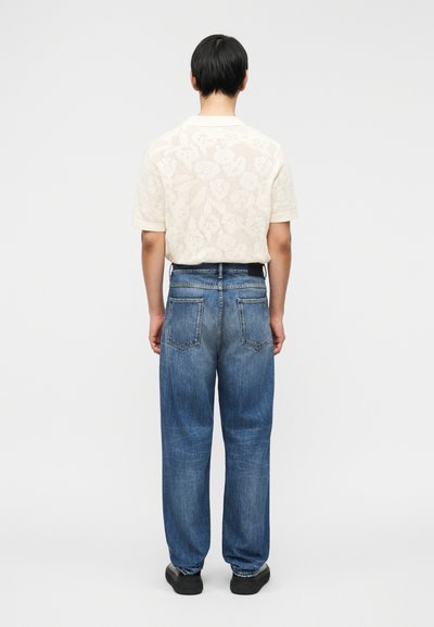 Ελαφρώς μπεζ πλεκτό κοντομάνικο πουκάμισο με λουλουδένιο σχέδιο, σε συνδυασμό με φαρδιά μπλε τζιν παντελόνια, εμφανιζόμενο από πίσω σε απλό φόντο.