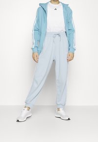Sweatshirt à capuche bleu clair avec des rayures blanches, associé à un pantalon de jogging bleu clair avec des rayures blanches sur les côtés, et des baskets blanches.