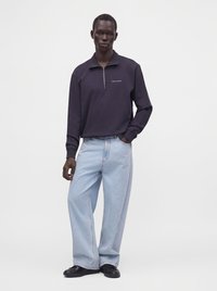 Άνδρας που φοράει μπλε sweatshirt με φερμουάρ μέχρι τη μέση και κείμενο, και ανοιχτό μπλε φαρδιά τζιν, στέκεται με το ένα χέρι στην τσέπη.