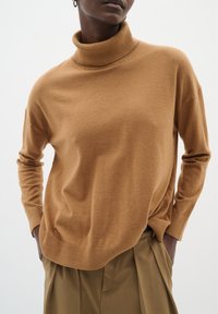 Brun rullekravepullover lavet af blødt stof, med lange ærmer, ribbet hals og kant og en afslappet pasform over højttaljede beige bukser.