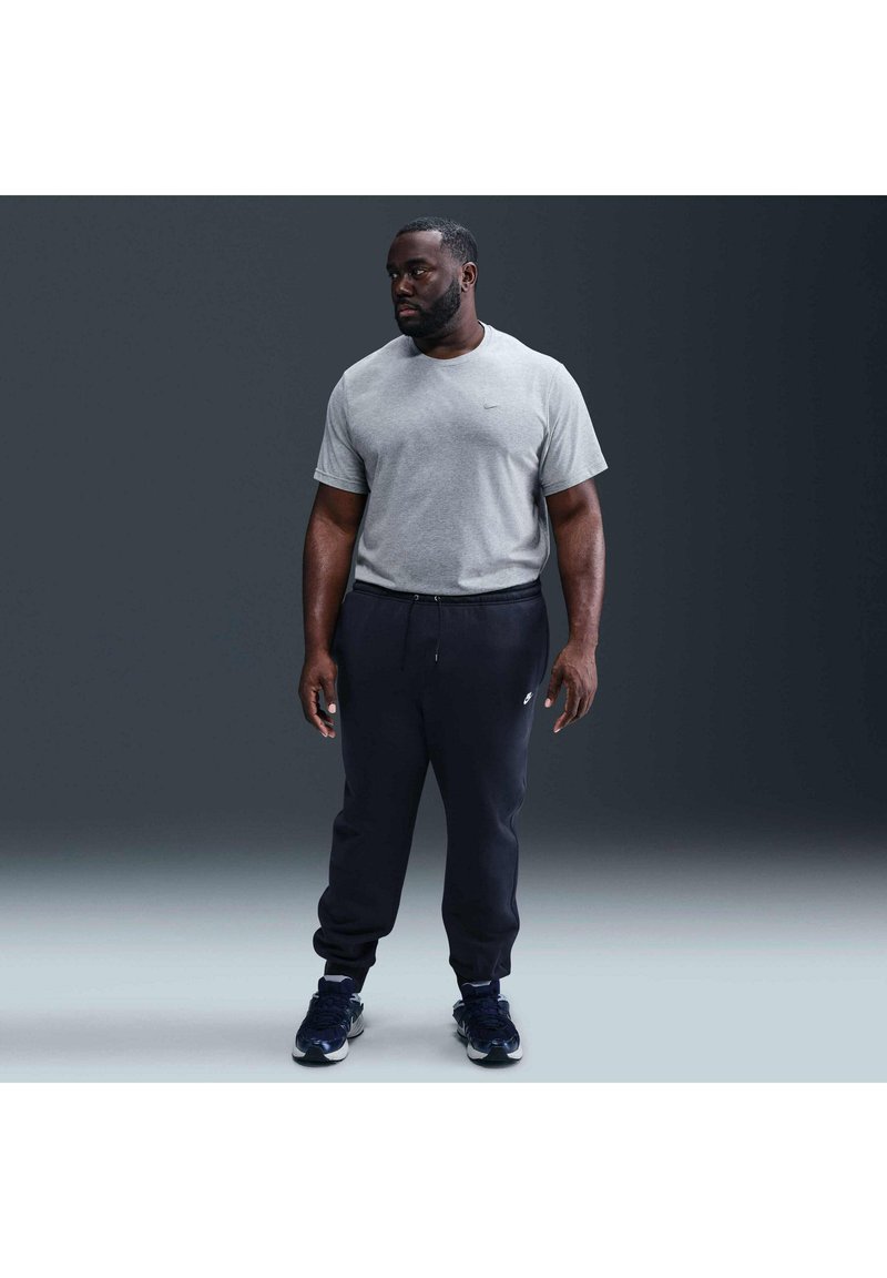 Uomo in piedi contro uno sfondo scuro, indossa una maglietta grigia a maniche corte, pantaloni della tuta neri e sneakers nere, guarda di lato.