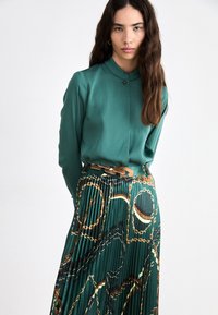 Grüne Bluse mit Kragen und Knopfdetails, kombiniert mit einem Faltenrock mit einem Kettenmuster in goldenen, braunen und dunkelgrünen Tönen.