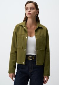 Veste à col en vert olive en matériau doux ; avec des boutons-pression et deux poches avant. Portée sur un haut blanc et un jean foncé.