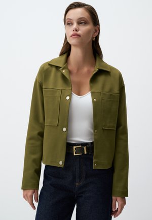Veste à col en vert olive en matériau doux ; avec des boutons-pression et deux poches avant. Portée sur un haut blanc et un jean foncé.