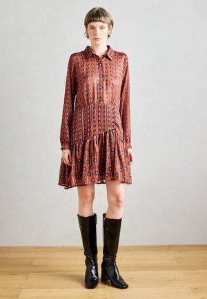 Dlhé rukávy, šaty po kolená so sťahovacím pásom, v hrdzavej farbe s modrými kvetinovými a geometrickými potlačami, skombinované s čiernymi lesklými čižmami po kolená.