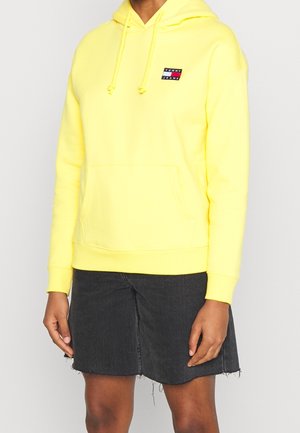 Person iført en klart gul Tommy Jeans hættetrøje med en frontlomme og sort frynset denimnederdel mod en ensfarvet baggrund.