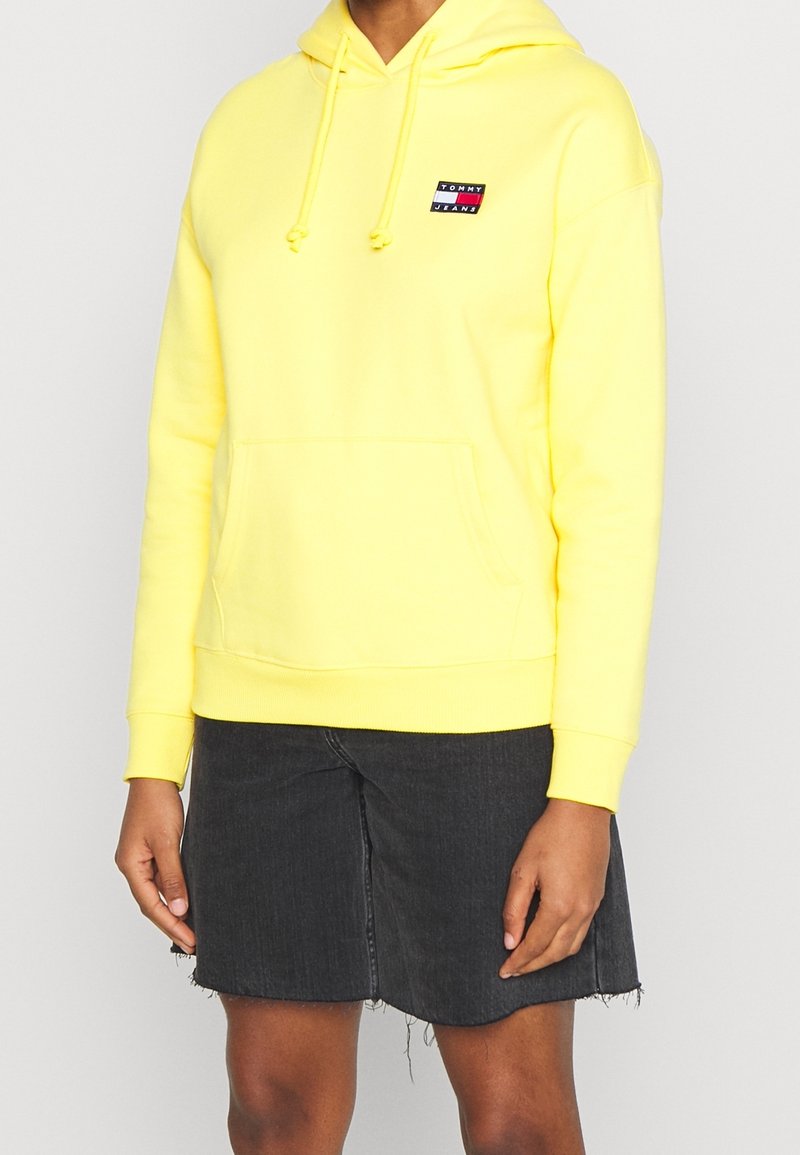Personne portant un sweat à capuche jaune vif Tommy Jeans avec une poche avant et une jupe en jean noire effilochée sur un fond uni.