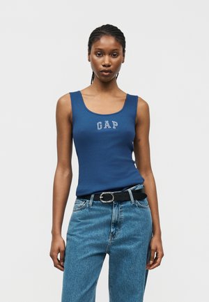 Fonott hajú nő kék GAP atlétafelsőben, világoskék farmernadrágban és fekete övvel, egy egyszerű fehér háttér előtt állva.