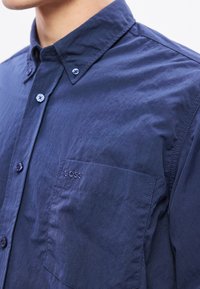 Cămașă bleumarin cu nasturi, realizată dintr-un material ușor, cu un buzunar pe piept, având o broderie subtilă cu logo și detalii contrastante la nasturi.