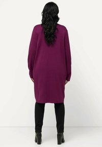Lila Strickkleid mit langen Ärmeln, leicht übergroßer Passform und gerippter Textur, kombiniert mit schwarzen engen Hosen und Stiefeln mit Absätzen.