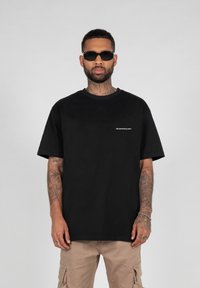 Schwarzes Baumwoll-T-Shirt mit Rundhalsausschnitt, kurzen Ärmeln und einem kleinen weißen Logo auf der linken Brust. Ein Paar rechteckige schwarze Sonnenbrillen wird getragen.