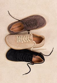 Tři semišové tenisky se šněrováním v hnědé, béžové a černé barvě, uspořádané překrývajícím se způsobem na béžovém texturovaném povrchu.