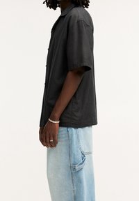 Camicia nera a maniche corte con vestibilità comoda e chiusura frontale con bottoni, indossata sopra pantaloni in denim azzurro chiaro, con tasca sull'anca e texture sbiadita.