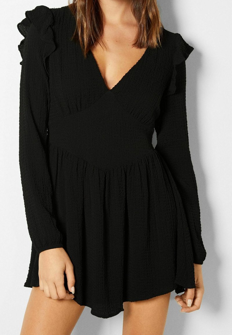 Robe noire à manches longues en tissu texturé, col en V et jupe évasée. Elle comporte des volants aux épaules et une taille cintrée.