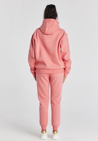 Persona de pie, de espaldas, vistiendo un sudadera rosa y pantalones de chándal a juego, con zapatillas blancas que tienen acentos dorados.
