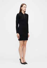 Γυναίκα που στέκεται, ντυμένη με εφαρμοστό μαύρο φόρεμα με μακριά μανίκια και φερμουάρ μπροστά, και μαύρες μυτερές ψηλοτάκουνες γόβες, μπροστά σε απλό λευκό φόντο.