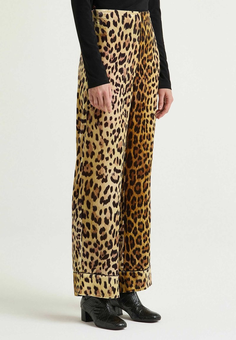 Pantaloni a gamba larga con stampa leopardata in tessuto morbido con diverse tonalità di marrone e beige, caratterizzati da un orlo ripiegato e indossati con scarpe nere.