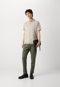Beige kötött pólóing rövid ujakkal, olajzöld, tapered szabású nadrággal párosítva. Fekete keresztpántos táska cipzárokkal és fekete bőr cipők.