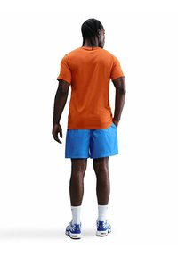 Conjunto atlético que incluye una camiseta corta naranja y pantalones cortos azules, combinados con zapatillas blancas y azules, sobre un fondo blanco.